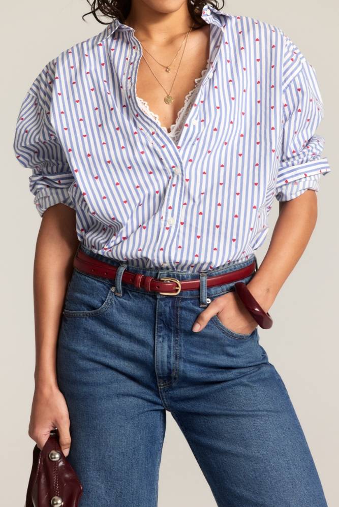 Kvinne med en stripete skjorte stukket ned i høytlivs blå jeans med rødt belte, stående med én hånd i lommen.