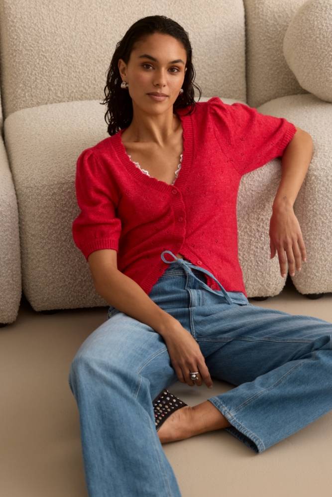 Kvinne i en rød bluse og blå jeans med vide ben som sitter avslappet ved siden av en beige sofa.