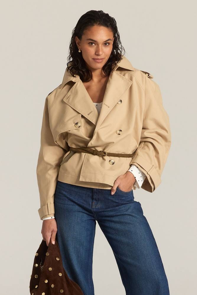 Kvinne i en kort beige trenchjakke stylet med blå jeans, som holder en brun håndveske.