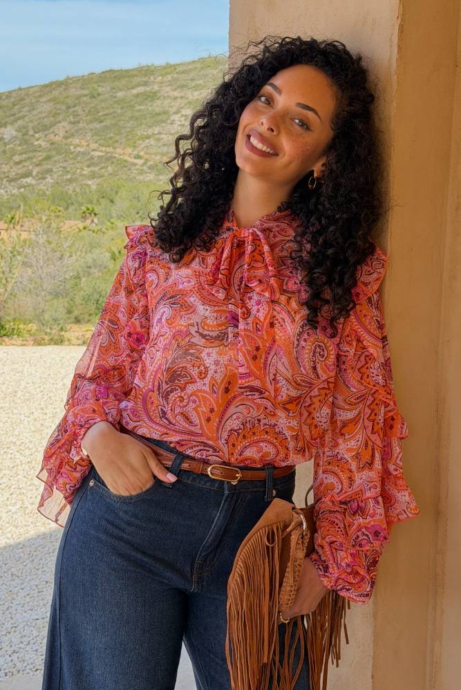 Kvinne i rosa paisleymønstret bluse med volangdetaljer og mørke jeans som lener seg mot en vegg utendørs.