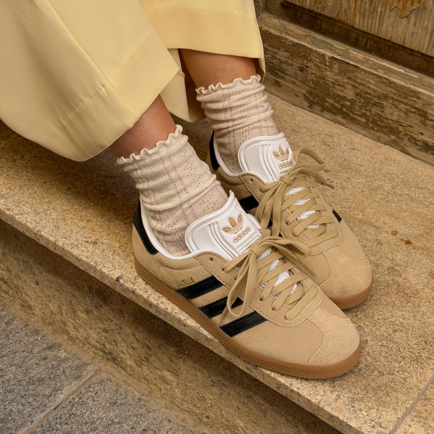 Lähikuva beigeistä Adidas-tennareista mustilla raidoilla, yhdistettynä röyhelöreunaisiin kermanvärisiin sukkiin ja keltaisiin housuihin kivisillä portailla.