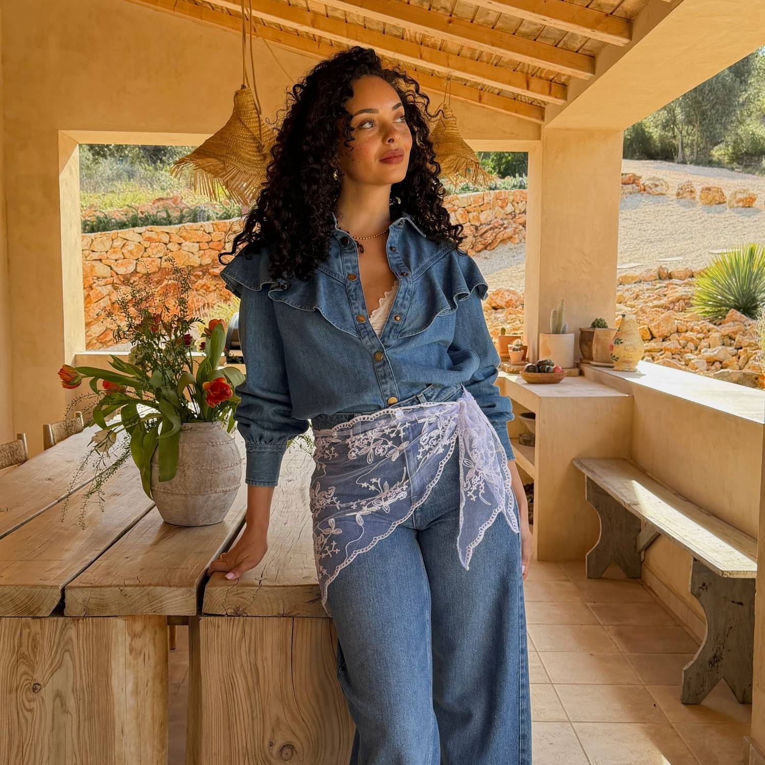 Kvinde i denimoutfit med flæsedetaljer, stående ved et træbord i en solrig udendørs setting med rustik indretning.