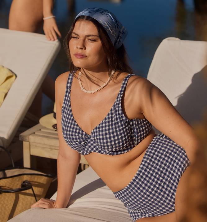 Kvinde i en blå bikini med ginghamtern sidder på en solseng ved poolen i sollys.