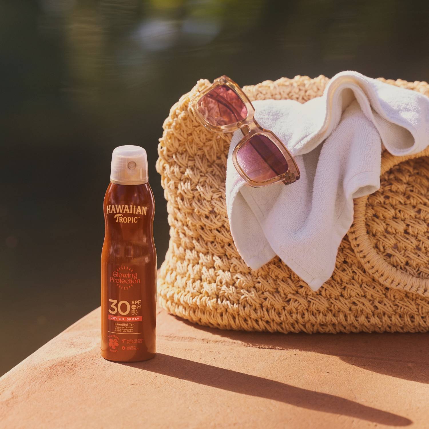 Flettet strandtaske i strå med solbriller og et håndklæde ved siden af en flaske solcreme på en solbelyst overflade.