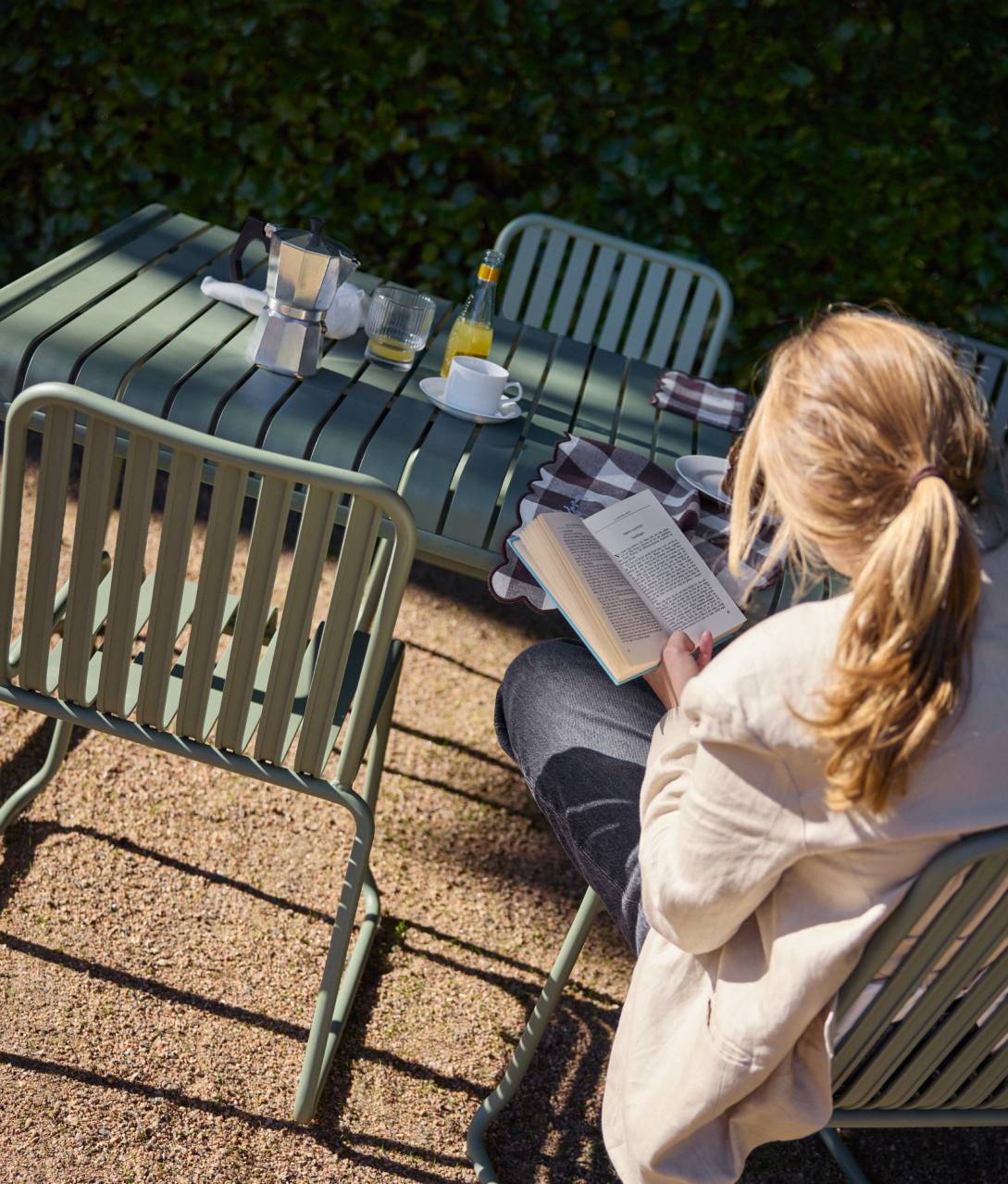 Nainen istuu ulkotuolilla ulkona pöydän ääressä lukemassa kirjaa.